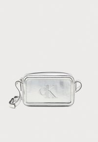 Strieborná metalická crossbody taška s embosovan�ým logom "CK", obdĺžnikový tvar, zipsové uzavretie a nastaviteľný popruh. Hladká textúra a elegantný dizajn.