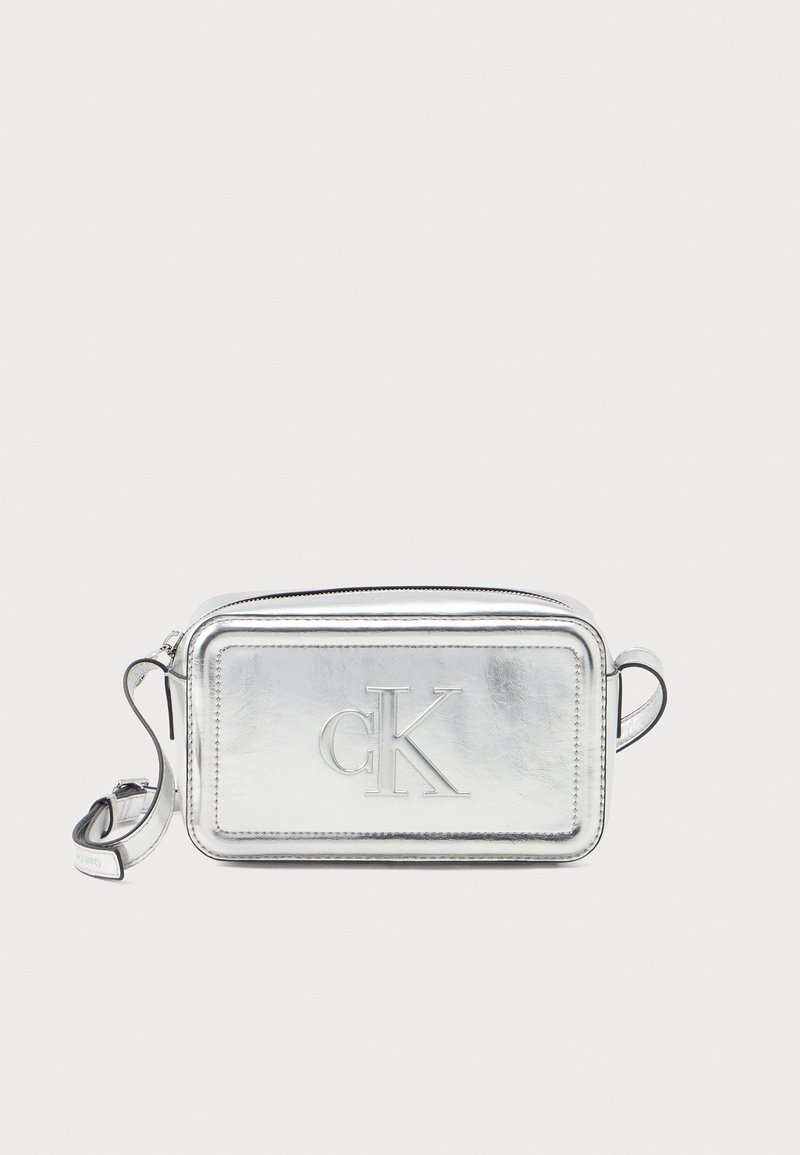 Strieborná metalická crossbody taška s embosovaným logom "CK", obdĺžnikový tvar, zipsové uzavretie a nastaviteľný popruh. Hladká textúra a elegantný dizajn.
