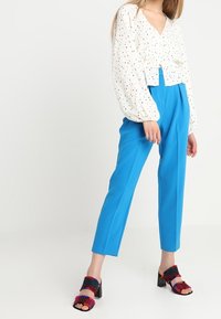 Blusa bianca con pois, maniche a sbuffo e scollatura a V, abbinata a pantaloni su misura blu brillante e sandali con tacco a blocco multicolori.