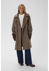 Jauna moteris šypsosi, dėvi rudą vilnonį paltą ant balto džemperio, tamsiai mėlynas platėjusias džinsines kelnes, rudus pirštines ir rudus batus.