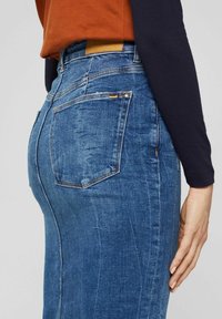 Modrá džínová sukně má přiléhavý střih, střední délku a viditelné zadní kapsy. Povrch je hladký s mírným vyblednutím.