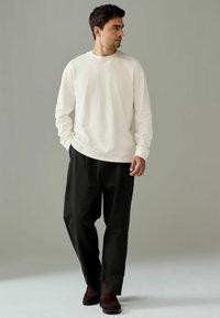 Long-sleeve cream shirt with a round neck, paired with loose-fitting black pants and brown shoes, standing on a light background.