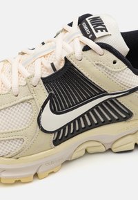 Nike Vomero 5 tenisice s bež mrežastim gornjim dijelom, crnim naglascima i svijetlim vezicama. Sadrži teksturirane panele i dizajn s podstavljenim potplatom.