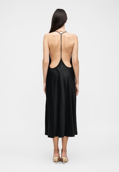 Γυναίκα που φοράει ένα μαύρο σατέν midi φόρεμα χωρίς πλάτη με λεπτές τιράντες και γυναικεία ψηλά πέδιλα σε nude απόχρωση, στέκεται μπροστά σε λευκό φόντο.