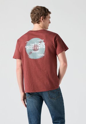 Jonge man van achteren, gekleed in een rood Levi's T-shirt met ronde grafische tekst en een blauwe spijkerbroek tegen een effen achtergrond.