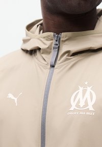 Μπεζ μπουφάν με φερμουάρ στο μπροστινό μέρος, λογότυπο Puma στο αριστερό στήθος και έμβλημα της ποδοσφαιρικής ομάδας της Μασσαλίας με το μότο "Droit Au But" στο δεξί στήθος.