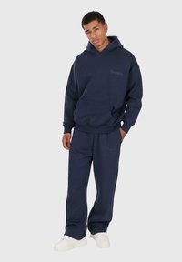 Junger Mann, der einen dunkelblauen Kapuzenpullover und passende Sweatpants mit dezenten "Dropsice" Logos trägt, steht mit einer Hand in der Tasche und weißen Sneakers.