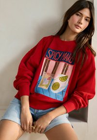 Raudona maikutė su dideliu grafikų dizainu, kuriame yra "SPICY MARG" tekstas, spalvingų gėrimų iliustracijos ir laisvas siluetas. Su džinsiniais šortais.