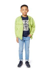 Veste zippée vert clair, t-shirt bleu marine avec imprimé palmiers, jeans bleu clair et chaussures de sport foncées sur un enfant se tenant debout devant un fond blanc.