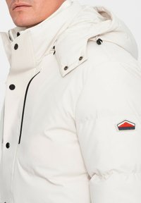 Giacca a vento bianca con colletto alto, chiusura a zip e bottoni a pressione, cappuccio regolabile, dettagli neri e un patch con logo sulla spalla sinistra.