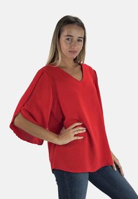 Rode V-hals top met brede vloeiende mouwen, textuurstof en een losse pasvorm, gecombineerd met donkere jeans voor een casual look.
