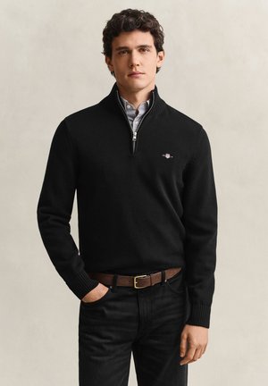 Jonge man die een zwarte trui met rits draagt over een geruit overhemd, zwarte jeans en een bruine riem, staand met één hand in de zak.
