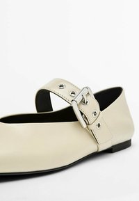 Lys beige flad lædersko med en bred strop, der har sølvhardware og øjestikdetaljer. Rund tå og lav, sort sål.