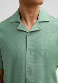 Camicia a maniche corte in materiale verde chiaro testurizzato con colletto dritto, cinque bottoni e una superficie liscia e tessuta.
