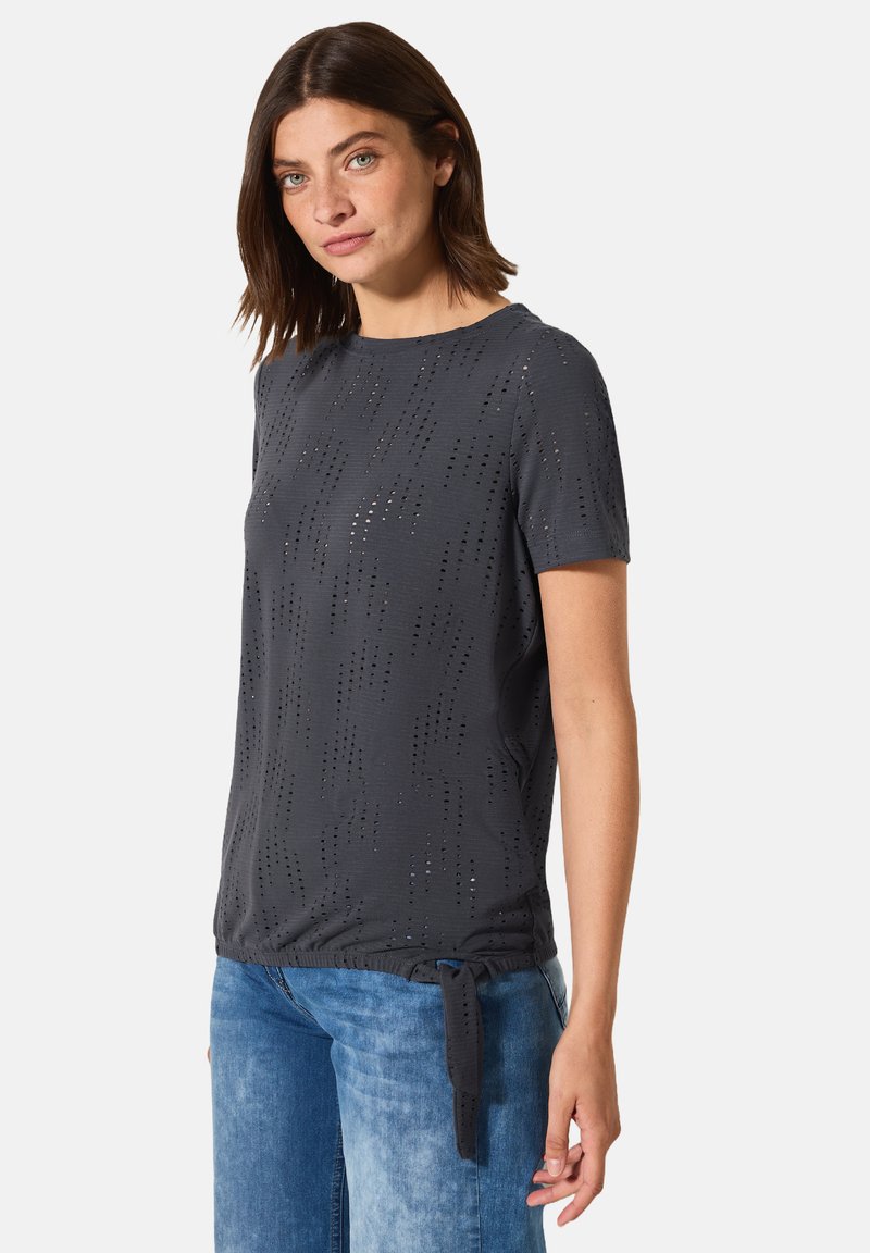 Haut gris à manches courtes avec un motif texturé et de petites découpes ; présente un nœud sur le côté à l'ourlet pour un ajustement réglable. Associé à un jean en denim clair.
