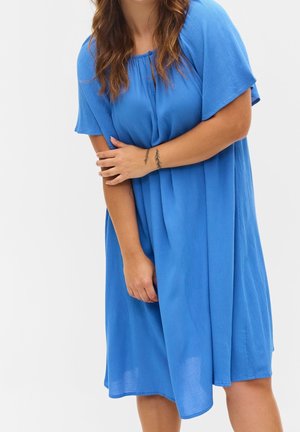 Frau mit langem braunem Haar, die ein lockeres, knielanges blaues Kleid trägt und mit einem Arm über dem anderen Handgelenk steht, auf dem kleine Tattoos zu sehen sind.