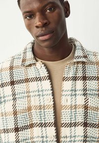 Jeune homme portant une chemise beige et une veste à carreaux boutonnée avec des motifs blancs, bruns et bleu clair, regardant légèrement sur le côté.