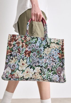 Person holder en stor rektangulær floral gobelintaske med burgunderrøde håndtag, iført lyst tøj og hvide sokker.