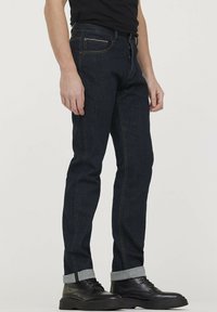 Jeans en denim bleu foncé avec une coupe slim, dotés d'une fermeture éclair, de poches avant et d'ourlets retroussés. Les accents incluent des coutures dorées et des éléments métalliques.