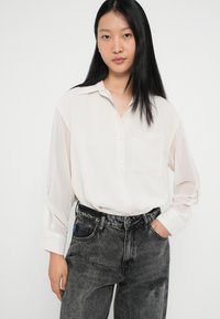 Light beige button-up blouse with a collar, long sleeves, and a chest pocket, paired with dark gray, high-waisted jeans.