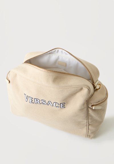 Μπεζ καμβάς τσάντα Versace με χρυσά φερμουάρ, μπροστινή τσέπη και το κεντρικό διαμέρισμα ανοιχτό που δείχνει λευκή επένδυση εσωτερικά.