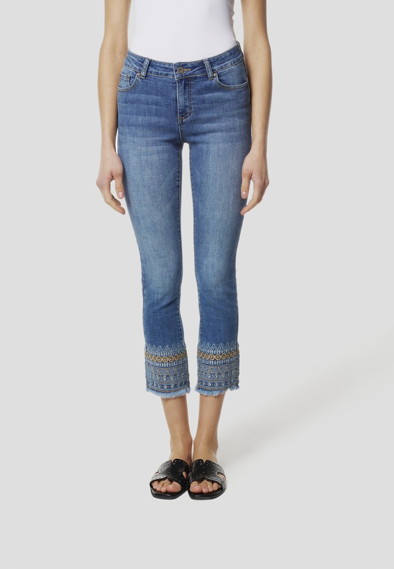Lichte blauwe denim capri-jeans met ingewikkelde geborduurde zoom met metallic accenten en een rafelige rand, gecombineerd met zwarte sandalen.