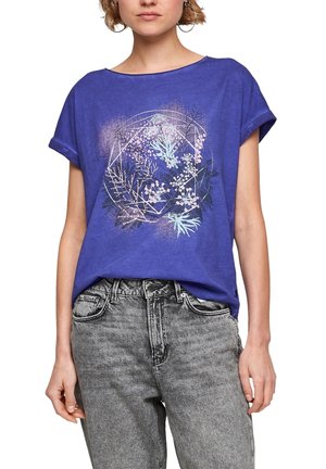 Femme portant un T-shirt bleu aux manches retroussées avec un motif floral géométrique pastel, associé à un jean gris clair taille haute.