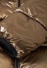 Nærbillede af en brun, blank, quiltet jakkeærme med sort elastisk manchet og synlig sort lynlås på puffet stof.