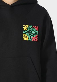 Sudadera negra con capucha que presenta un gráfico cuadrado con formas de plantas en verde, amarillo y rojo. El material es suave y tiene un bolsillo delantero.
