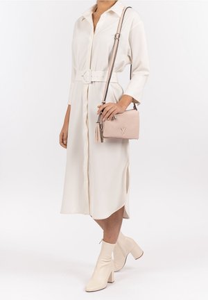 Vrouw in een witte midi-jurk met riem, crèmekleurige enkellaarsjes en een lichtroze crossbody tas met franjerand.