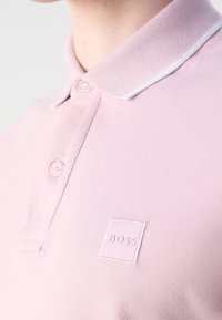 Polo rose clair avec deux boutons et un petit logo "BOSS" sur le côté gauche de la poitrine, porté par une personne.