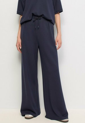 Persona que lleva pantalones anchos de pierna suelta de color azul oscuro con cintura ajustable y top de manga corta a juego en azul oscuro, de pie contra una pared lisa.
