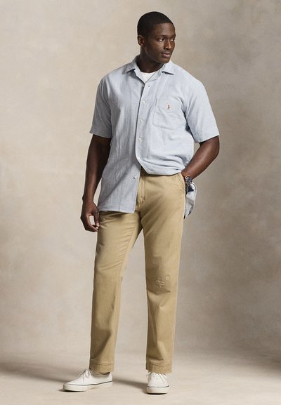 Moški, oblečen v svetlo modro srajco s kratkimi rokavi in gumbi, bež hlače, bele superge, stoji z eno roko v žepu, gleda v stran.