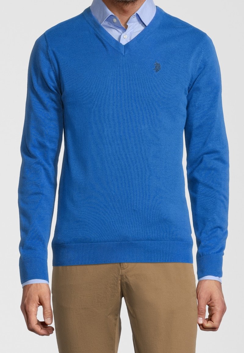 Blå, stickad v-ringad tröja med ribbstickade ärmslut och nederkant, prydd med en liten broderad logotyp på bröstet, bärs över en ljusblå skjorta med krage.