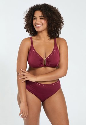 Uśmiechnięta kobieta z kręconymi włosami w bordowym dwuczęściowym kostiumie kąpielowym ze złotymi akcentami, stojąca ze skrzyżowanymi ramionami na jasnym tle.