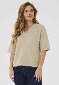 Pull en tricot beige avec un col en V, des manches courtes et une texture douce. Présente une coupe décontractée et une ourlet se terminant au-dessus de la taille.