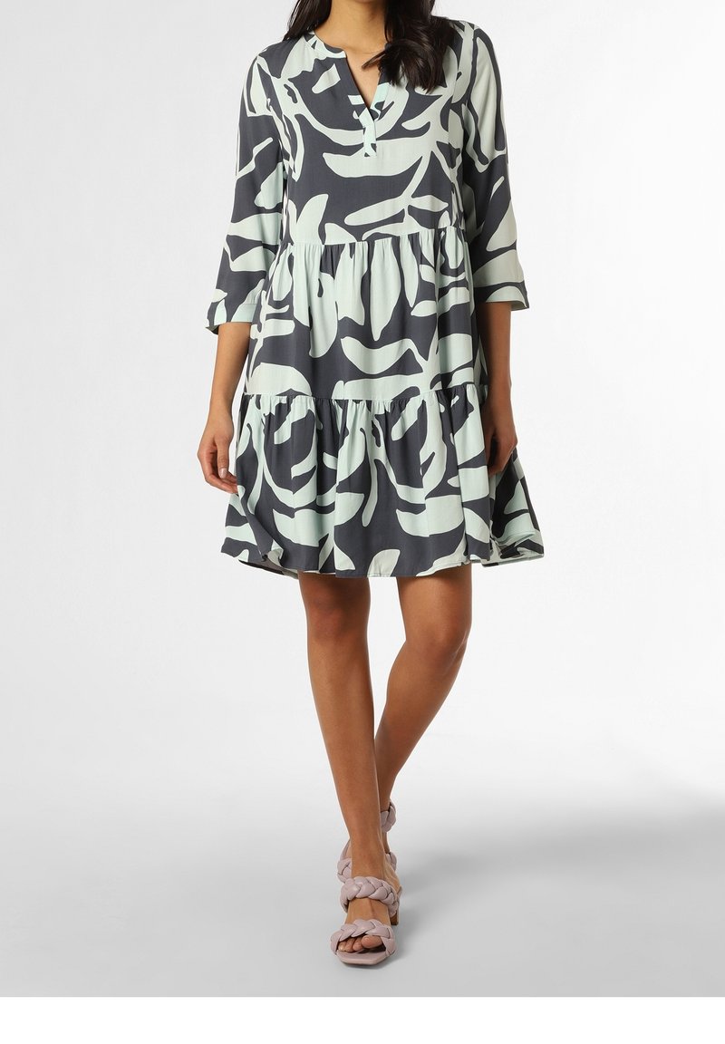 Femme portant une robe ample à étages avec des motifs abstraits de feuilles noires et vert pâle, manches trois-quarts, et sandales à brides tressées.