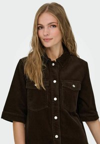 Brūna korduroja krekla jaka ar lielām sudraba krāsas pogām, krūšu kabatām un aizrullētām īsajām piedurknēm. Faktūras audums ar vertikālām rībām.