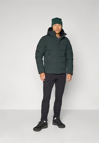 Tamsiai žalia paminkštinta striukė su kapišonu, užtrauktukais užsegamomis kišenėmis ir raštuotu paviršiumi. Dėvima su juodomis kelnėmis ir juoda avalyne.