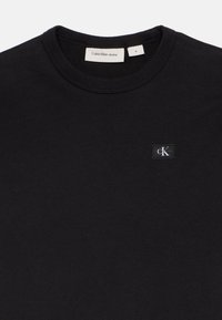 Juodas medvilnės džemperis su apvaliu kaklu, ant krūtinės esančiu mažų stačiakampiu Calvin Klein Jeans logotipo užsiuvimu. Lygus paviršius.