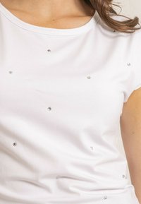 T-shirt blanc à manches courtes en tissu doux, décoré de petites strass dispersées. Texture lisse avec un col rond.