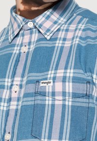 Niebieska koszula w kratę z jasnoróżowymi i białymi paskami, zapinana na guziki, z kieszonką na piersi i etykietą „Wrangler” na kieszeni.