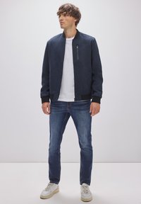 Laivaston pommikauppi, jossa on vetoketjullinen tasku, kiinteä pinta, ribattu hihansuukaulus, yhdistetty sinisiin farkkuihin ja valkoisiin lenkkareihin, neutraali t-paita alla.