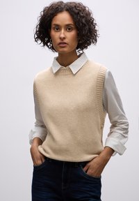 Beige gebreide mouwloze trui met ribgebreide accenten, gedragen over een witte blouse met kraag. Model met handen in de zakken, met donkere denim jeans.