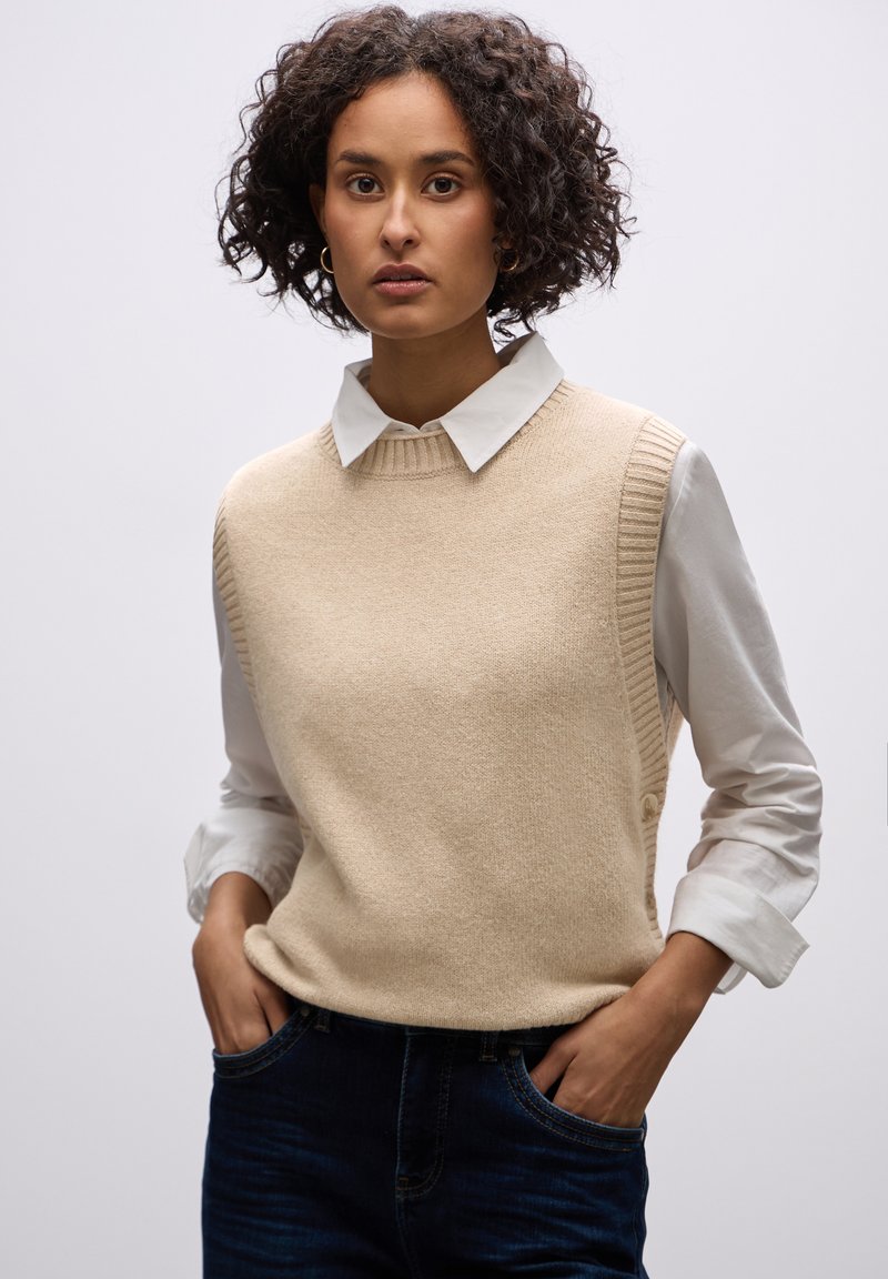 Beige gebreide mouwloze trui met ribgebreide accenten, gedragen over een witte blouse met kraag. Model met handen in de zakken, met donkere denim jeans.