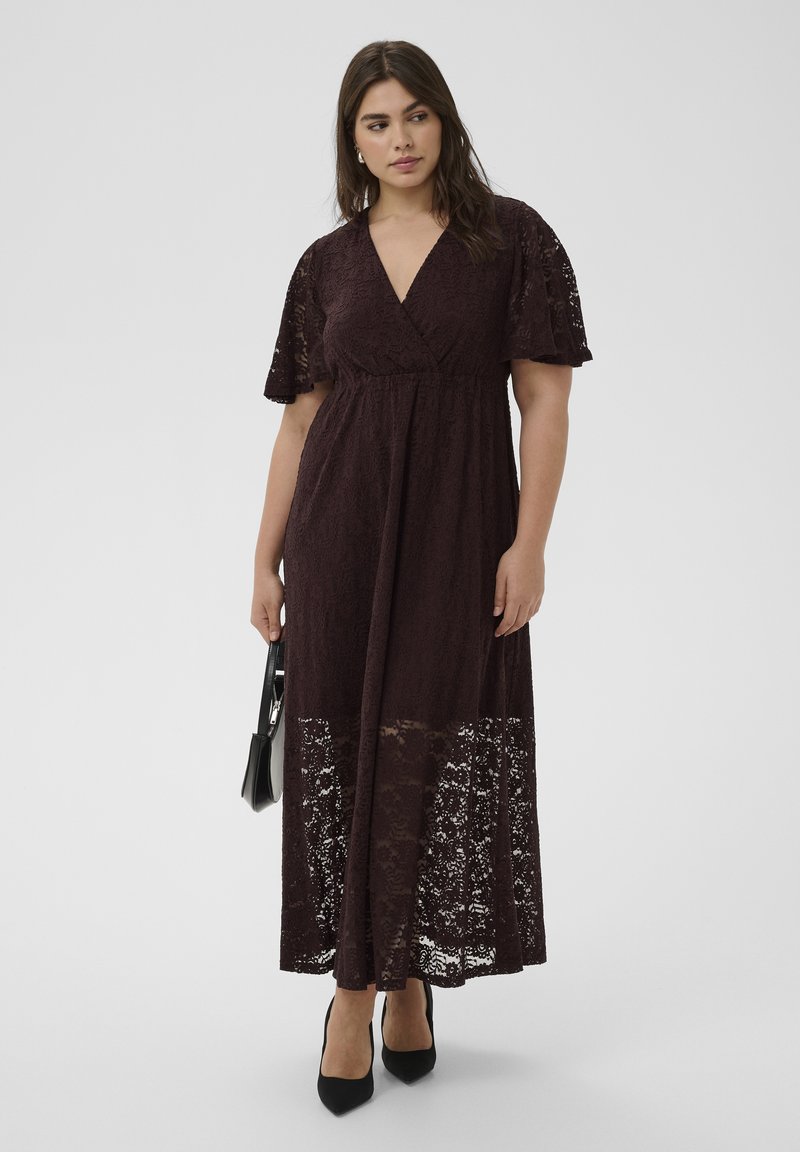 Femme portant une robe maxi en dentelle marron foncé avec des manches courtes volantées et un décolleté en V, tenant un sac à main noir, debout devant un fond uni.