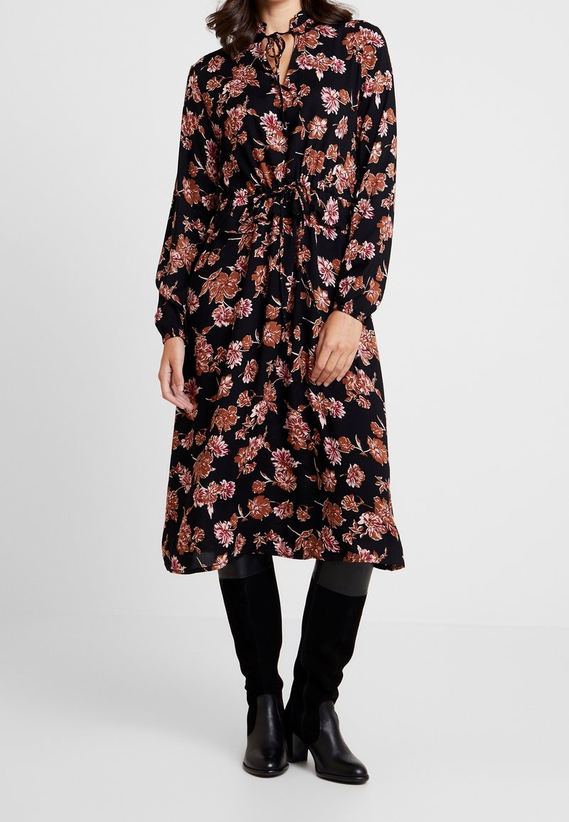 Femme portant une robe midi noire à imprimé floral marron, taille cintrée, manches longues, associée à des bottes hautes noires jusqu'aux genoux.