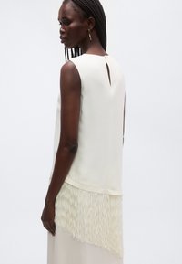 Top crem fără mâneci cu un decupaj în spate, având un detaliu texturat cu franjuri la tiv. Material neted cu o siluetă dreaptă.