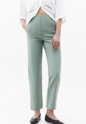 Vrouw draagt hoog-taile, rechte lichtgroene broek met verticale naden, een wit loszittend shirt en zwarte platte schoenen, staat met één hand in de zak.