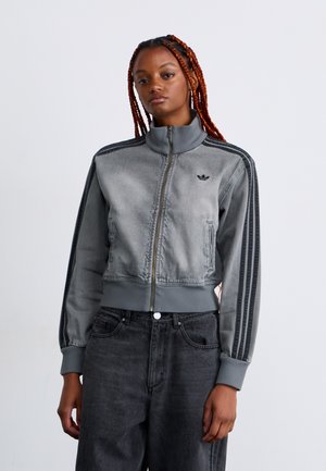 Junge Frau mit geflochtenem rotem Haar, die eine graue, geschnittene Zip-Jacke mit schwarzen Streifen und passenden dunkelgrauen, hochtaillierten Jeans trägt.